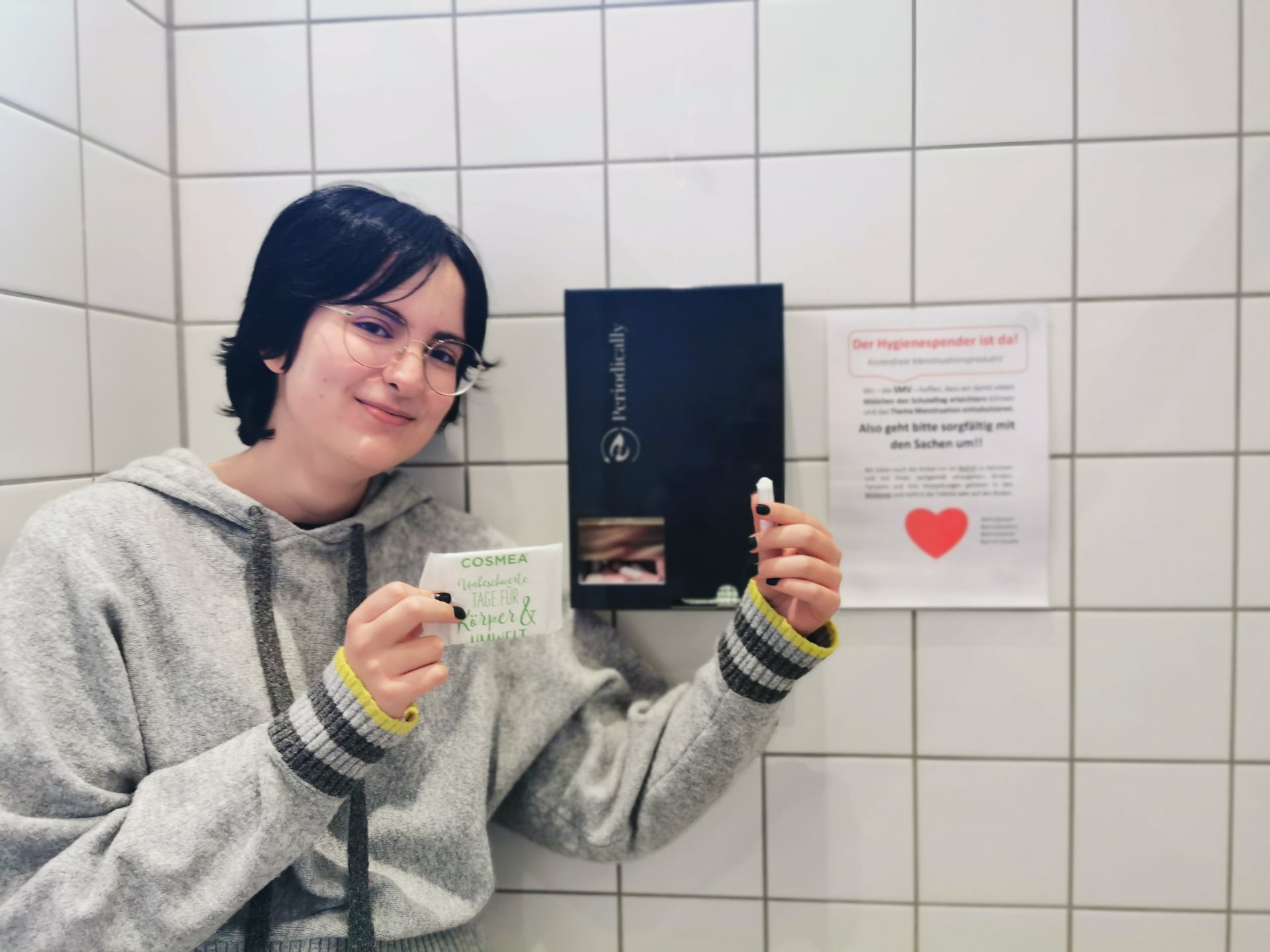 Person mit Brille vor gefliester Wand hält Bindenpackung und Tampon neben einem Hygienespender.