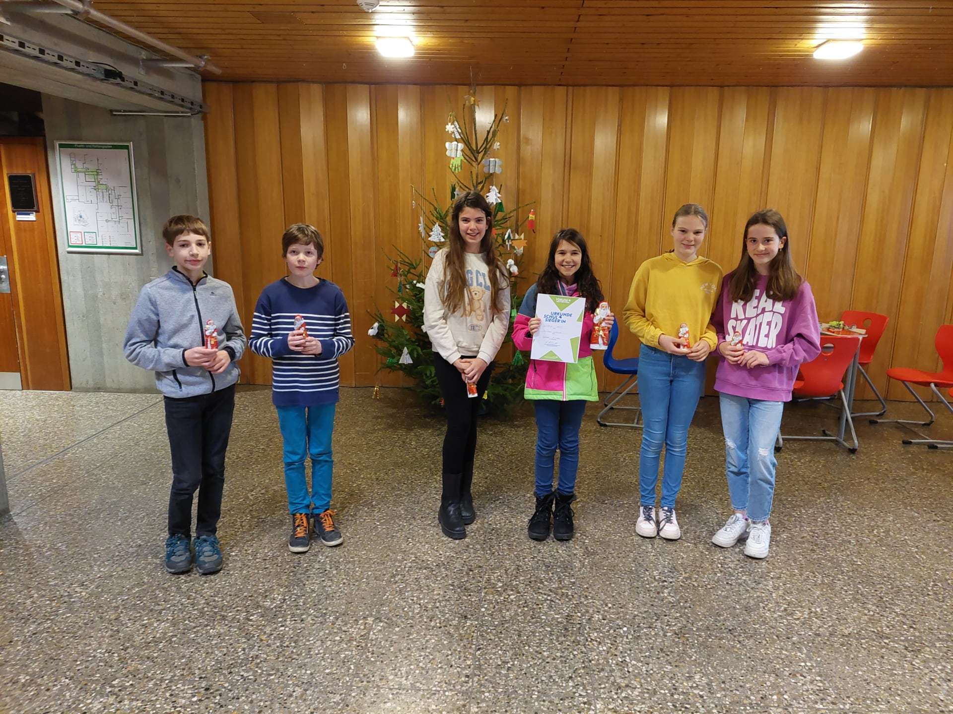 Sechs Kinder stehen nebeneinander vor geschmücktem Weihnachtsbaum, halten Schoko‑Nikoläuse; ein Kind zeigt Urkunde.