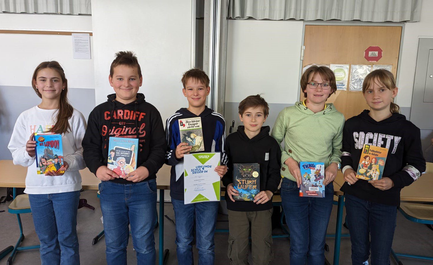 Sechs Schulkinder stehen in einer Reihe im Klassenzimmer, halten Bücher; mittleres Kind zeigt Urkunde.