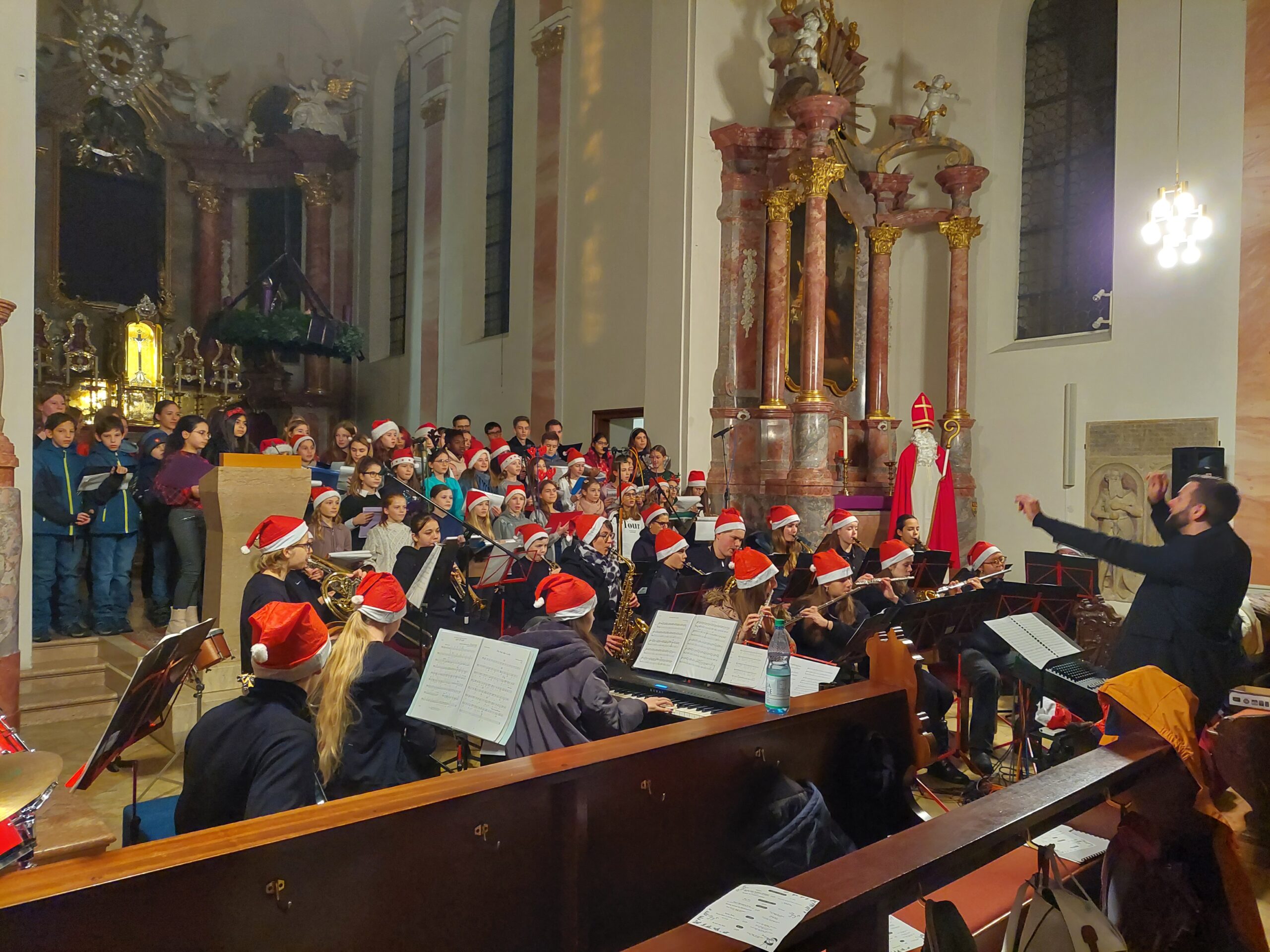 Jugendchor und Orchester in barocker Kirche, viele mit Weihnachtsmützen, Dirigent und Nikolausfigur
