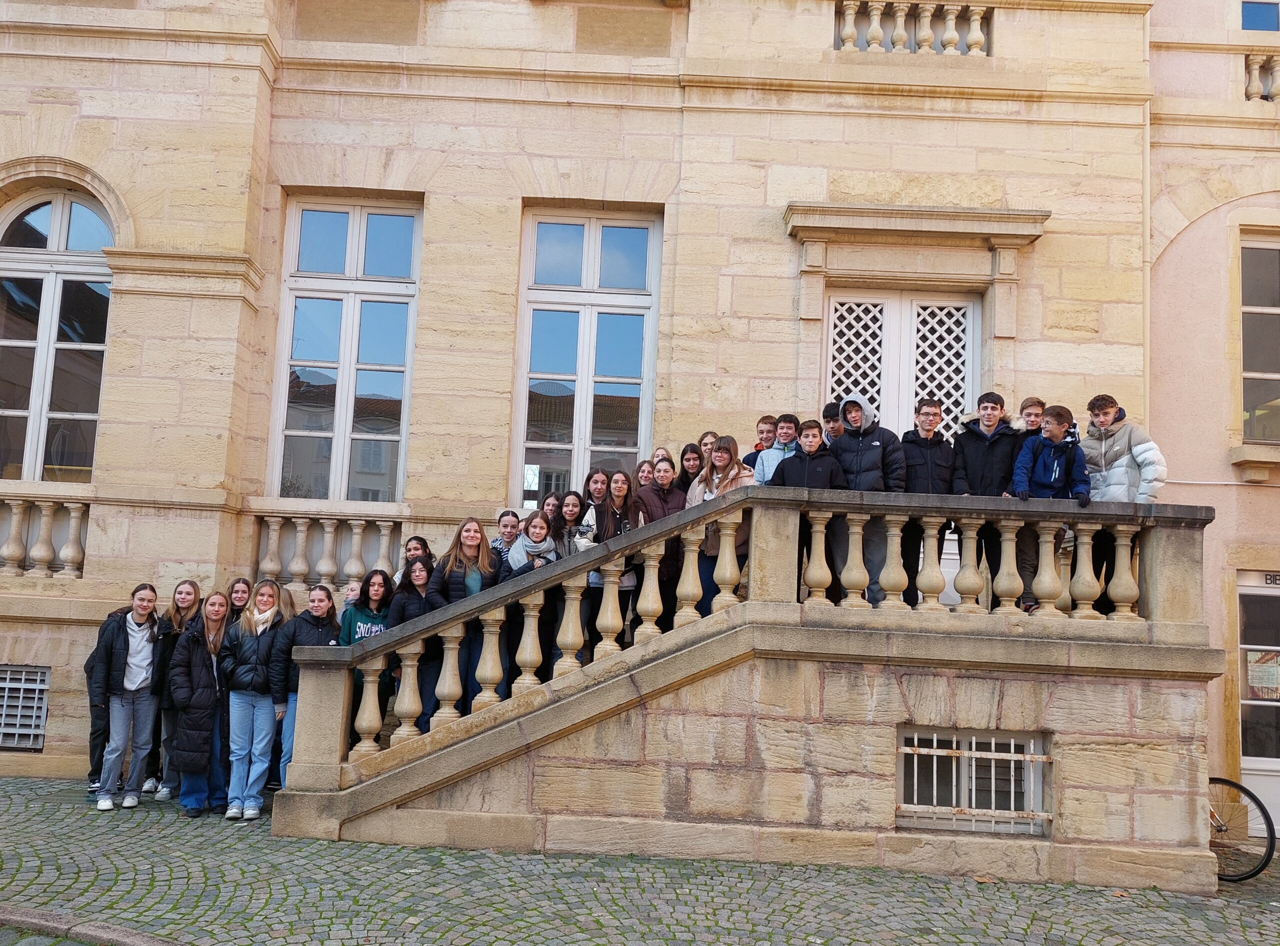 Schülergruppe auf steinerner Treppe vor historischer Sandsteinfassade mit großen Fenstern.