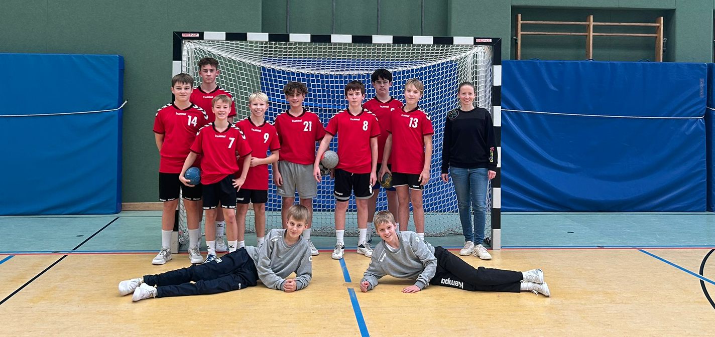 Jugend-Handballmannschaft in roten Trikots posiert vor Tor in Sporthalle, zwei Spieler liegen vorne.