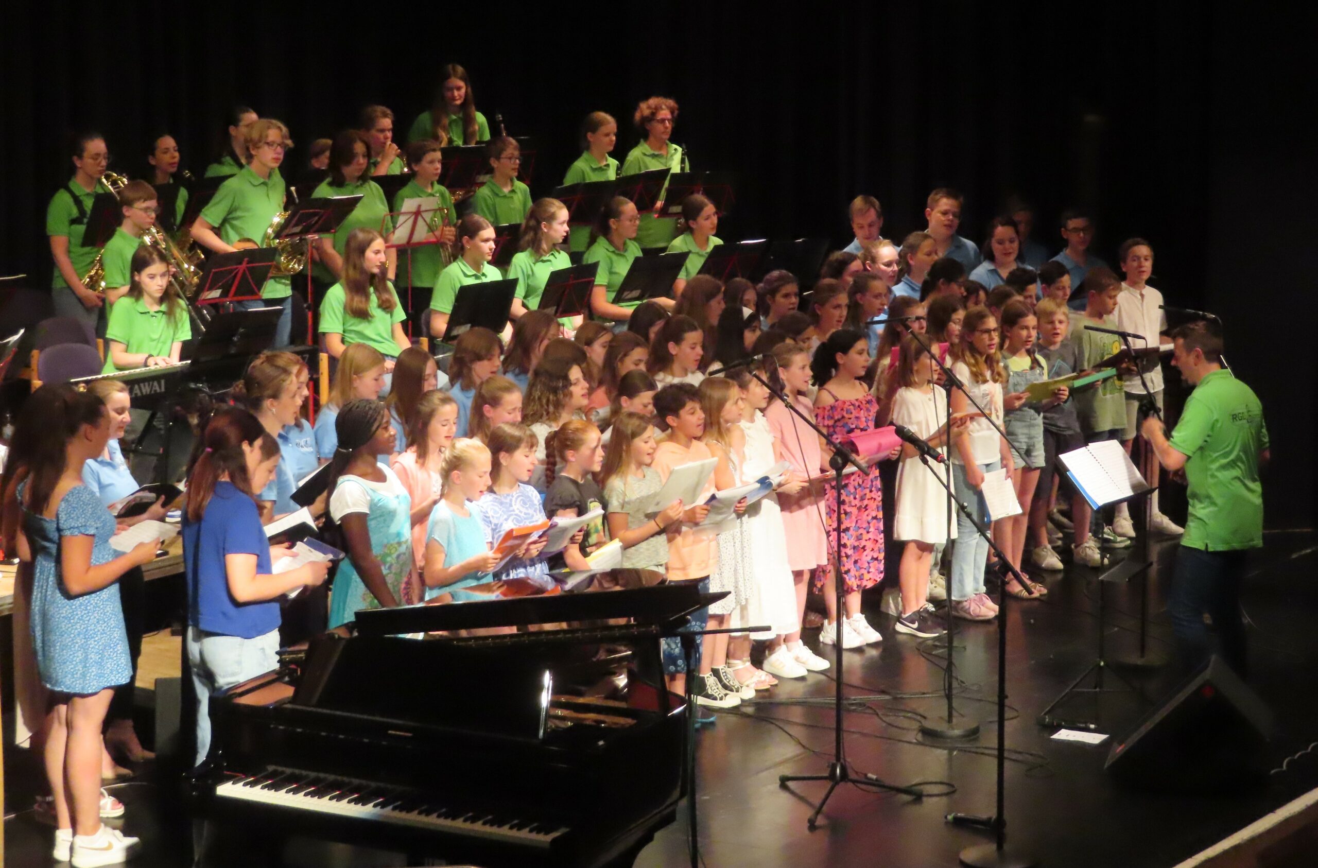 Kinder- und Jugendchor mit Schulorchester auf Bühne, singend vor Flügel; Dirigent in grünem Polo.