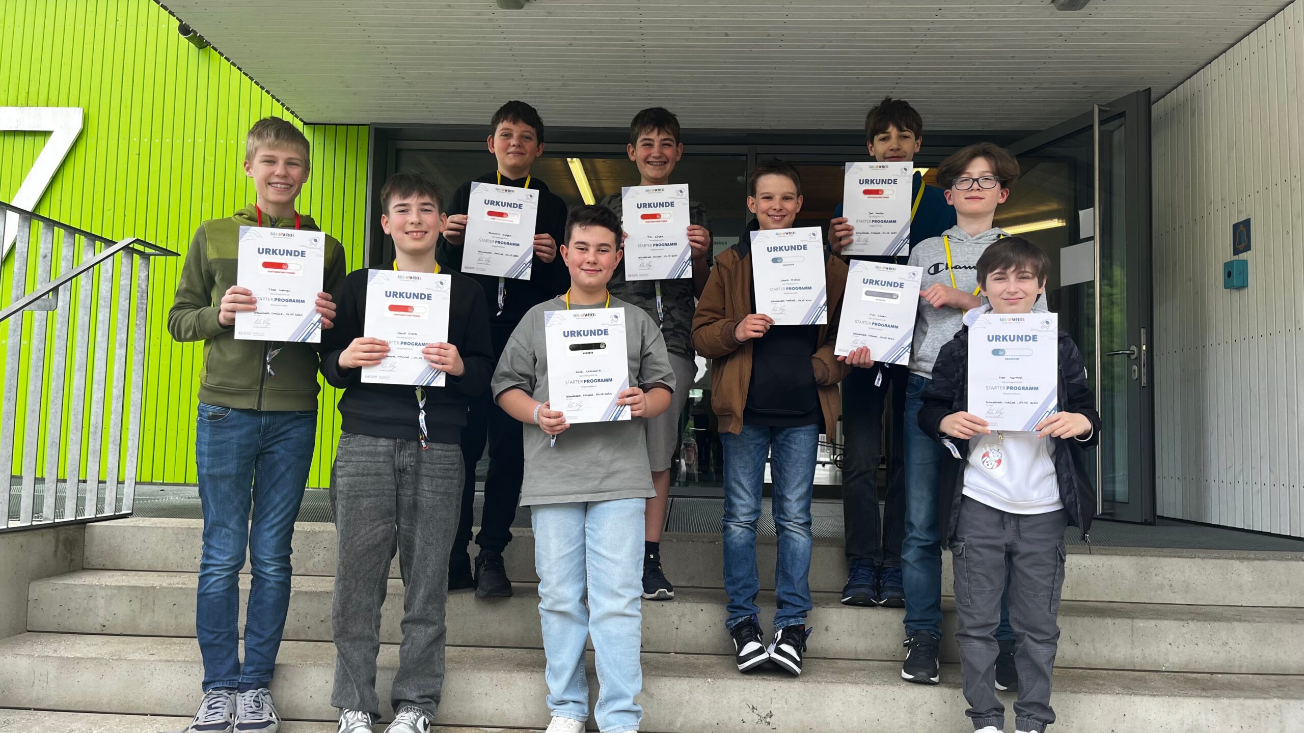 Zehn Jungen auf Treppe vor Schule, lächelnd, halten Urkunden und Medaillen.