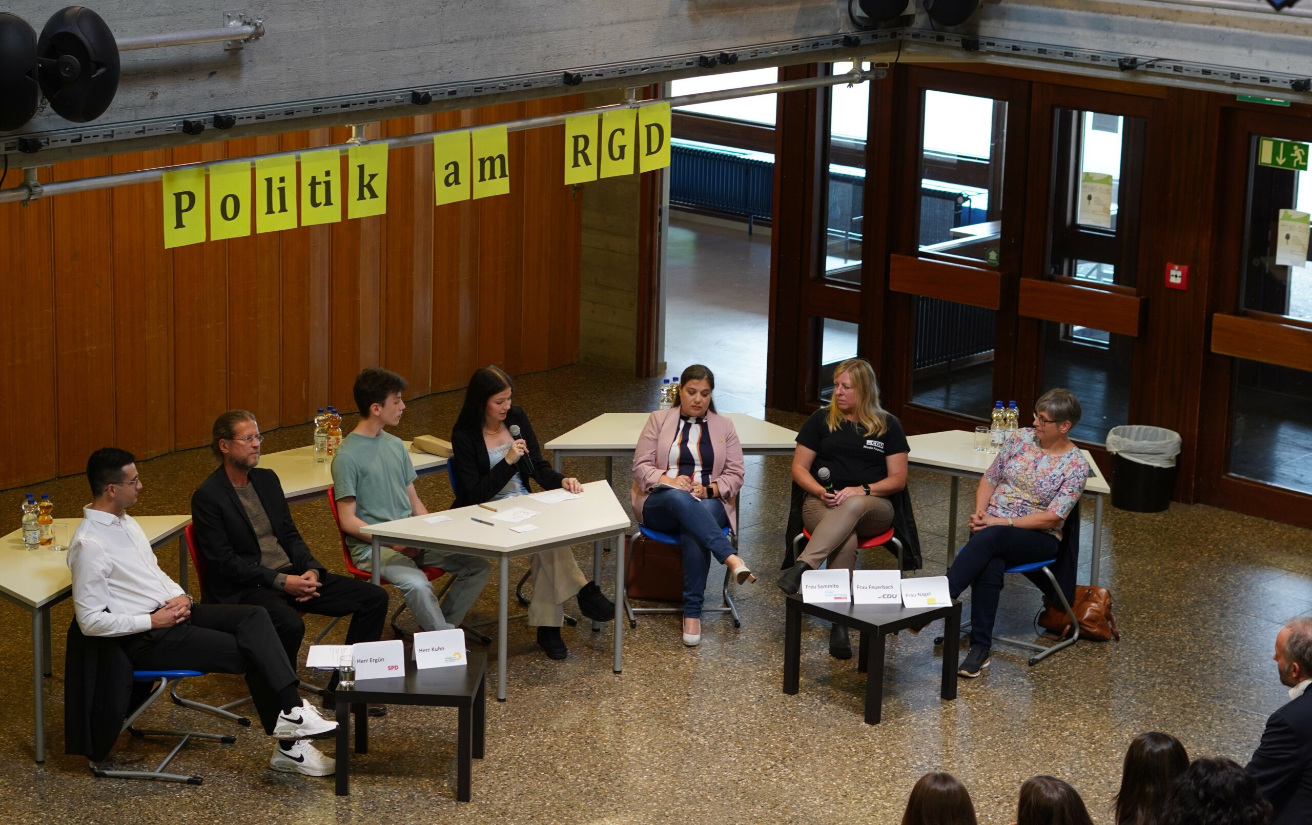 Podiumsdiskussion in Schulfoyer: sechs Teilnehmende sitzen an Tischen, gelbe Banner Politik am RGD