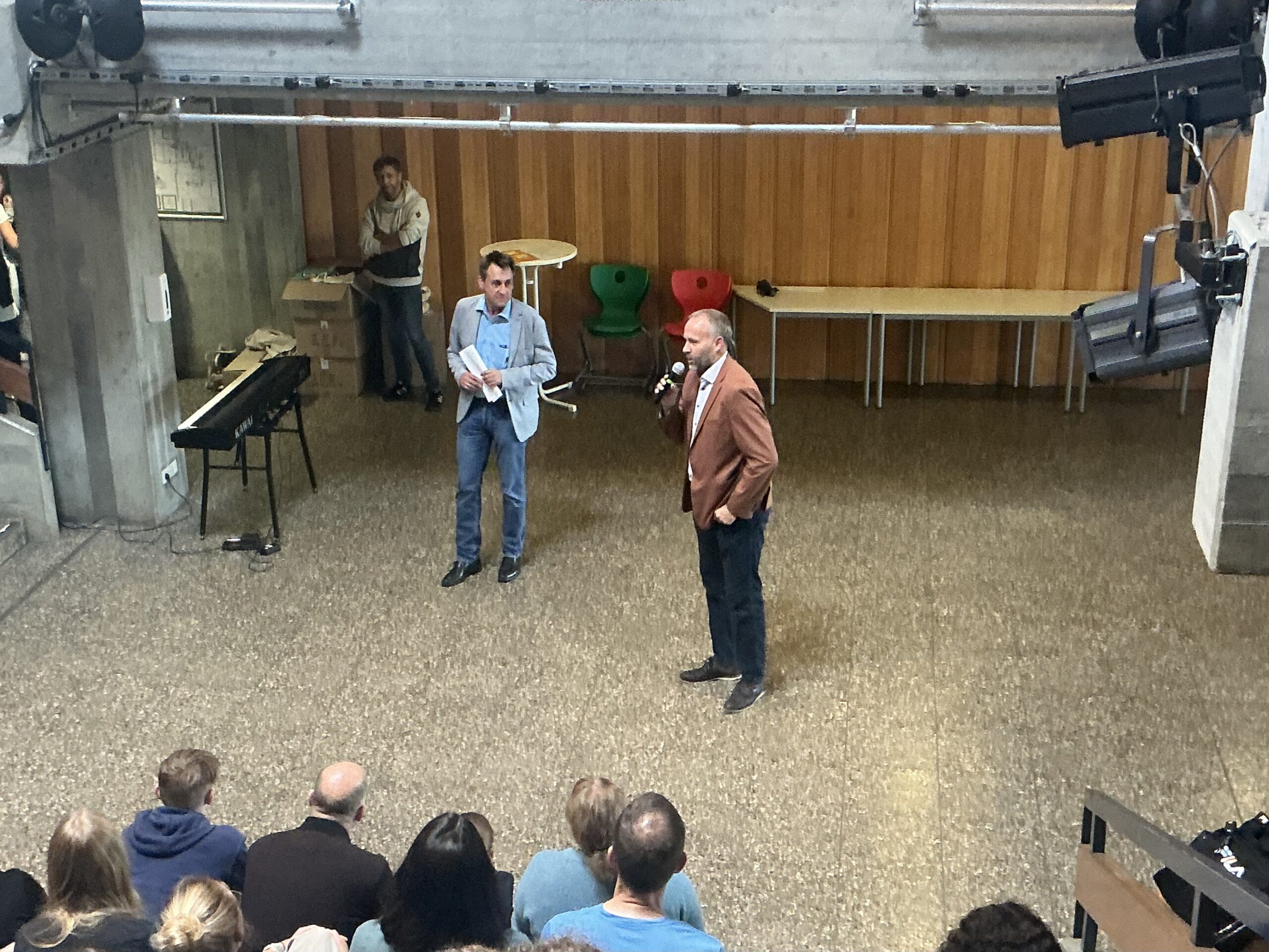 Blick von oben auf Publikum; zwei Männer vorne, einer spricht ins Mikrofon vor Holzpaneelwand.