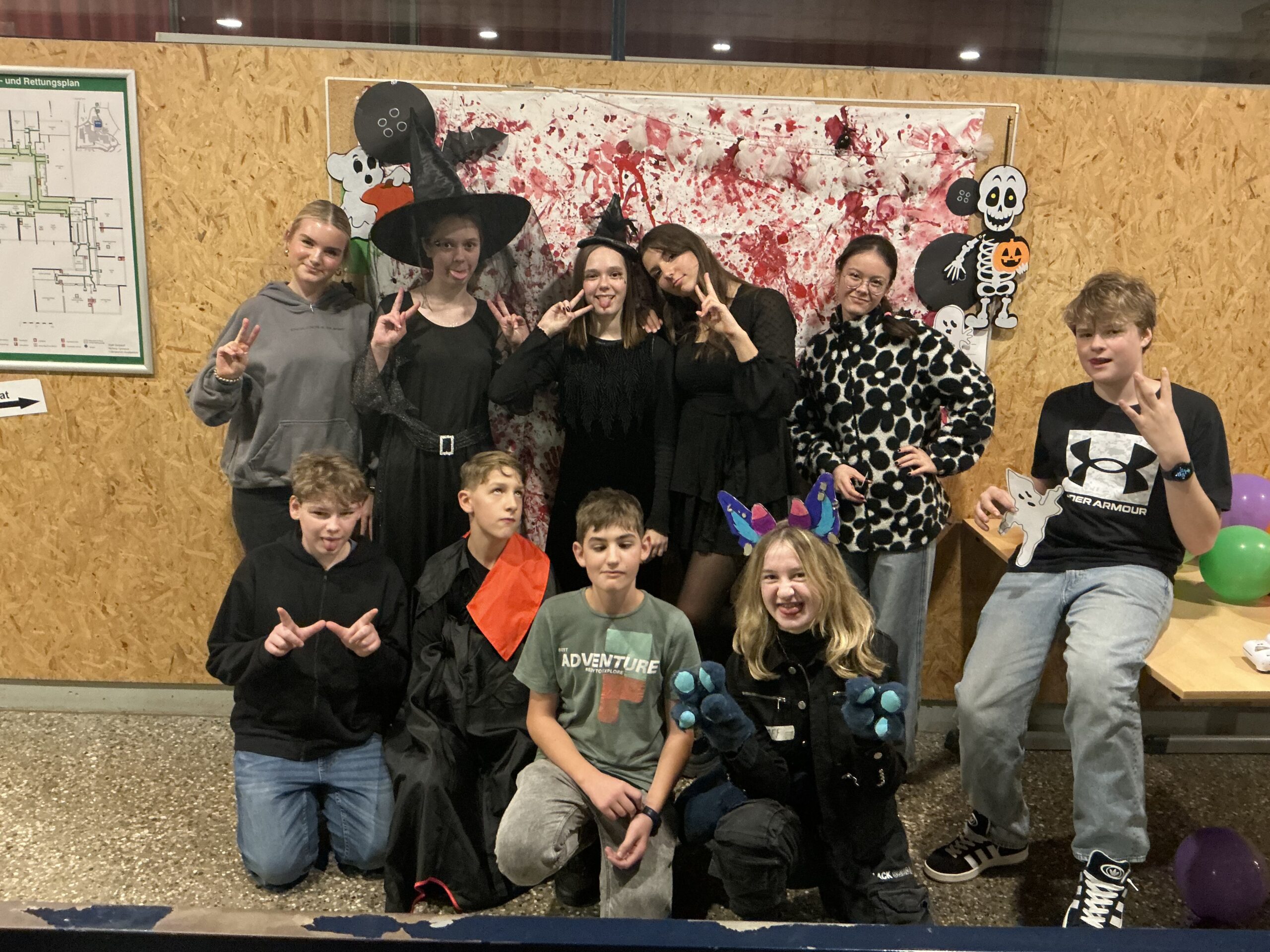 Gruppe Jugendlicher in Halloween-Kostümen (Hexen, Monster) posiert vor blutgespritztem Fotohintergrund und Skelett-Deko.