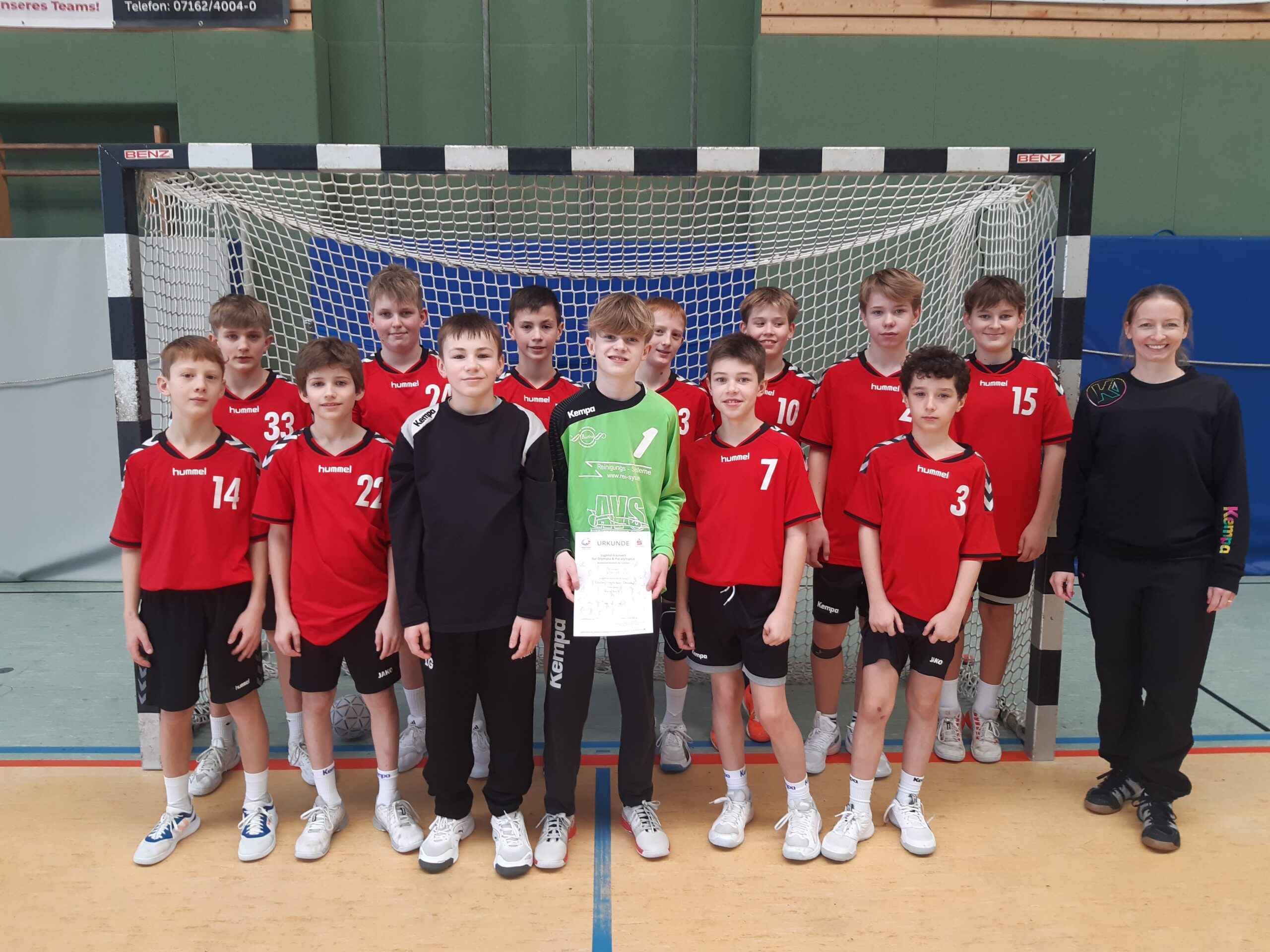 Jugend-Handballmannschaft in roten Trikots vor Tor, Torwart mit Urkunde, rechts lächelnde Trainerin.
