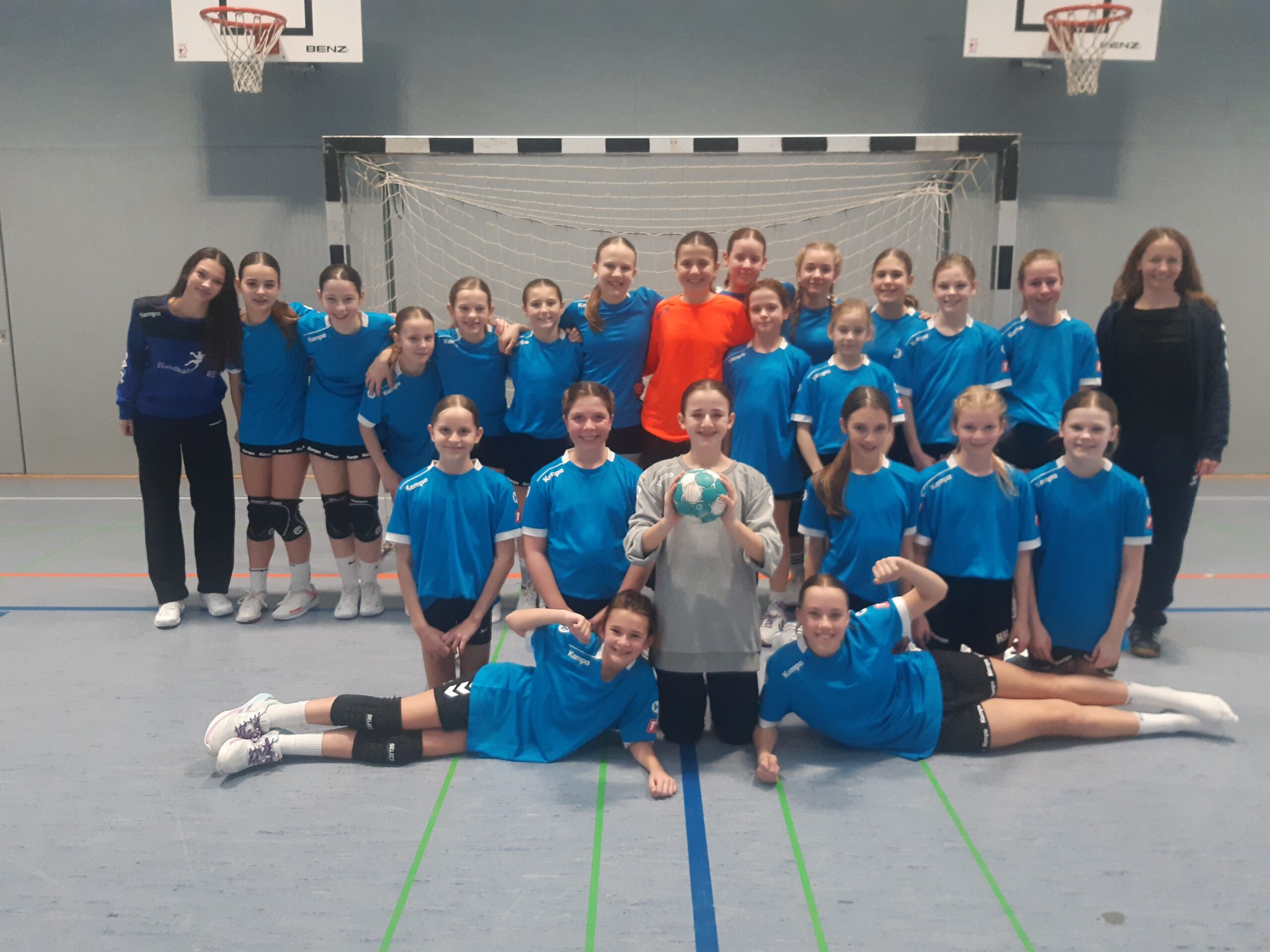 Jugend-Handballteam in blauen Trikots vor Tor in Sporthalle, Mädchen posieren, eines hält Ball, zwei Trainerinnen seitlich
