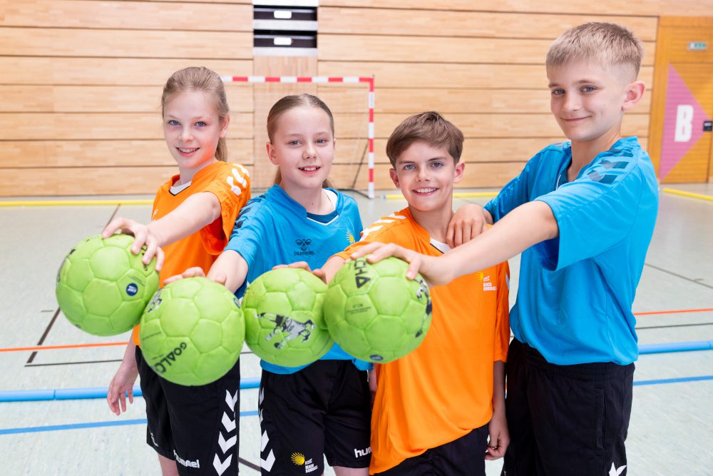 Vier Kinder in Sporthalle halten grüne Handbälle zur Kamera, lächelnd in orange-blauen Trikots vor Handballtor.