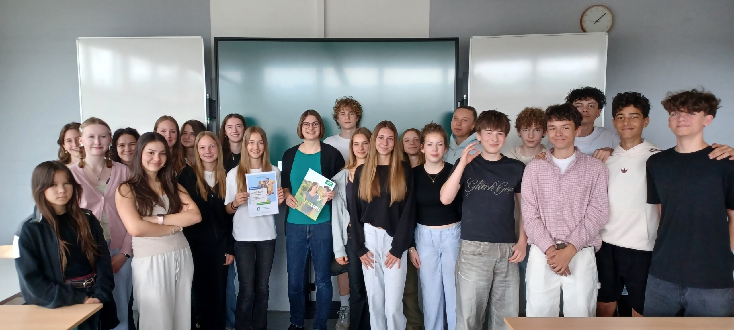 Schulklasse mit Lehrerin vor digitaler Tafel, zwei Schülerinnen halten Zertifikat und Broschüre.