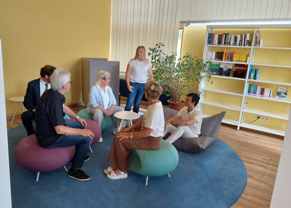 Sechs Erwachsene im Gespräch auf runden Sitzhockern in einer hellen Leseecke mit Bücherregal und Pflanze
