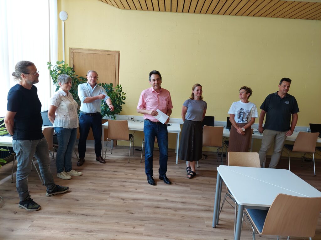 Sieben Personen stehen in einem hellen Besprechungsraum; der Mann in der Mitte hält ein Papier und spricht.