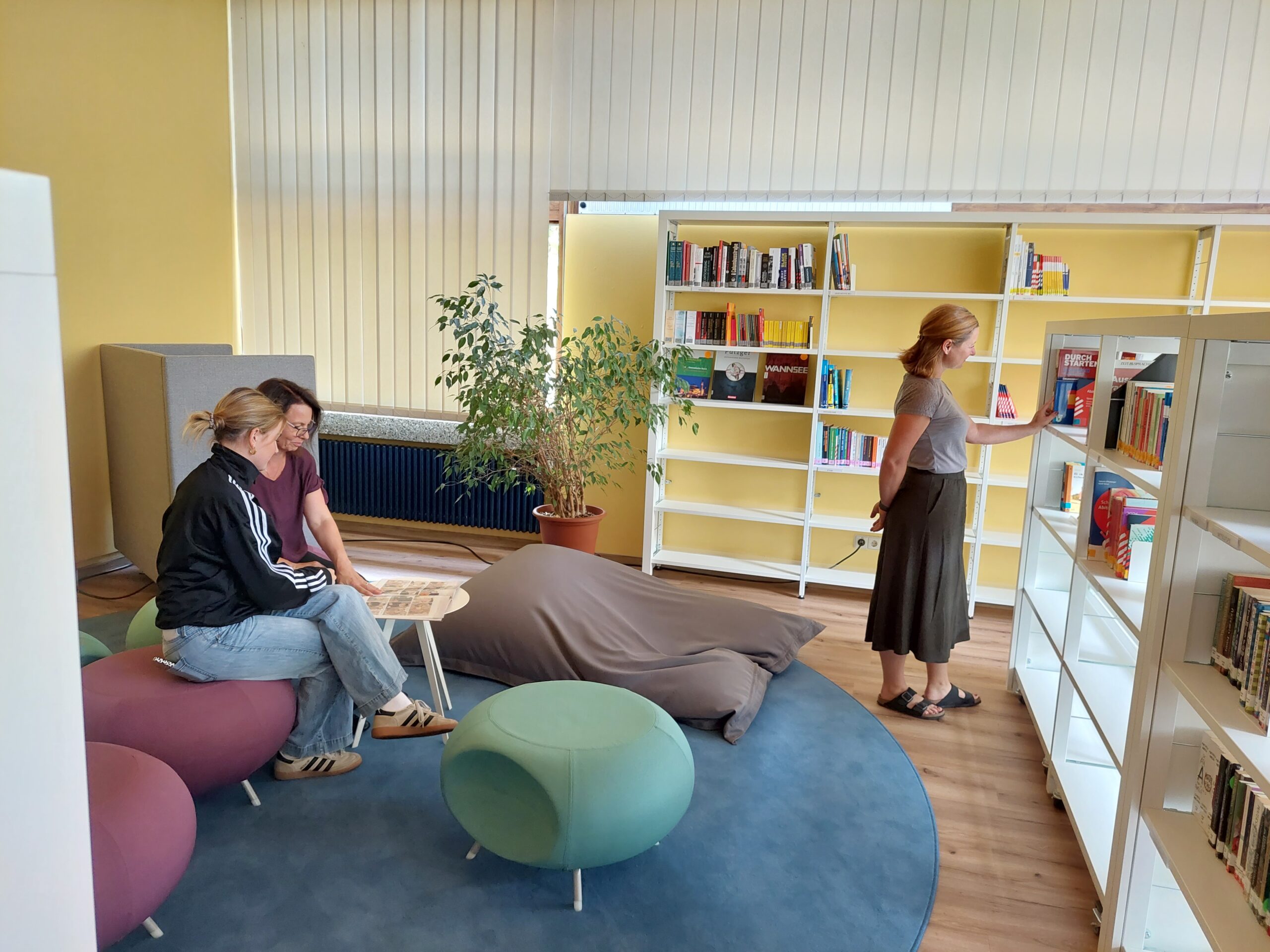 Lesebereich in Bibliothek: zwei Frauen sitzen mit Zeitung, eine Frau steht und sucht ein Buch an Regalen.
