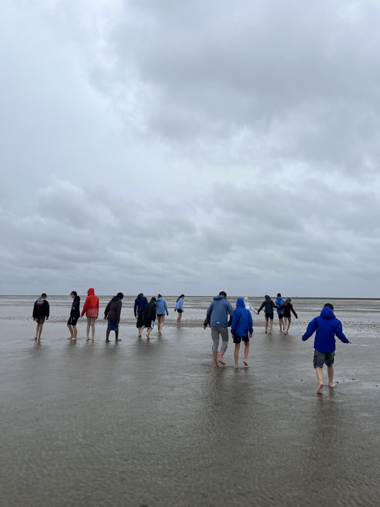 Gruppe Menschen wandert barfuß über nassen Wattboden Richtung Horizont unter grauem, wolkenverhangenem Himmel.