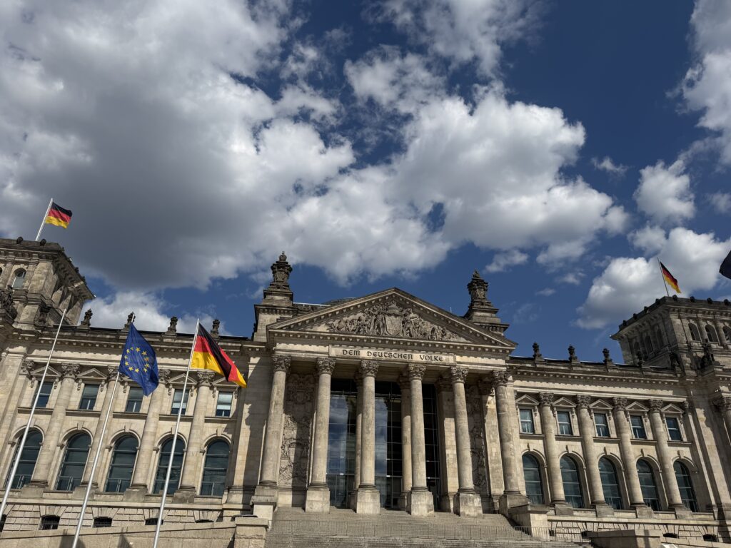 Reichstagsgebäude mit Säulen, der Inschrift 'Dem Deutschen Volke' und wehenden Deutschland‑ und EU‑Flaggen vor Wolkenhimmel.