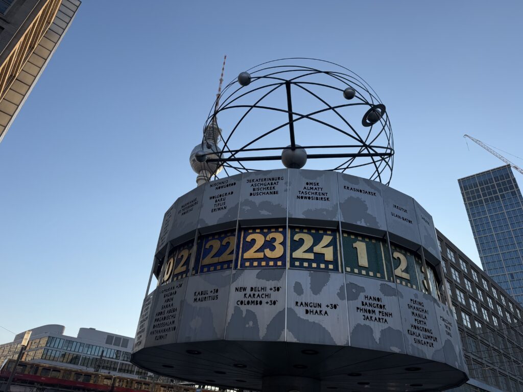 Weltzeituhr am Alexanderplatz mit Kugelgitter-Aufbau und Berliner Fernsehturm im Hintergrund unter blauem Himmel.