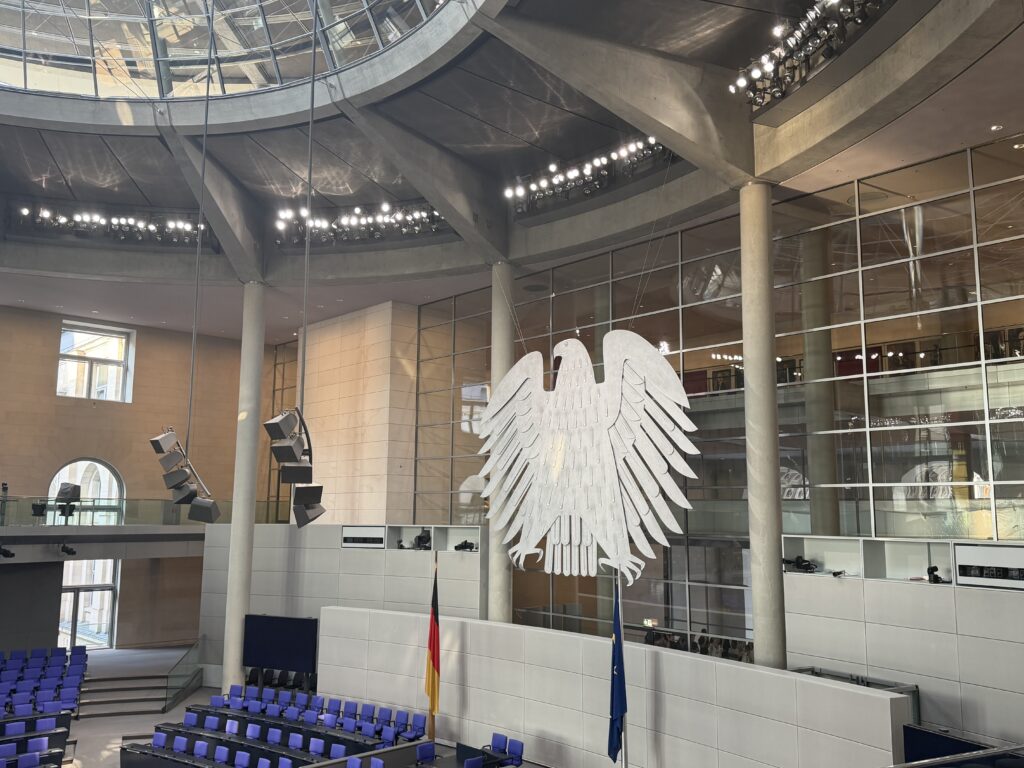 Plenarsaal mit hängendem Bundesadler, Reihen blauer Sitze, deutscher und EU‑Flagge vor großer Glasfassade