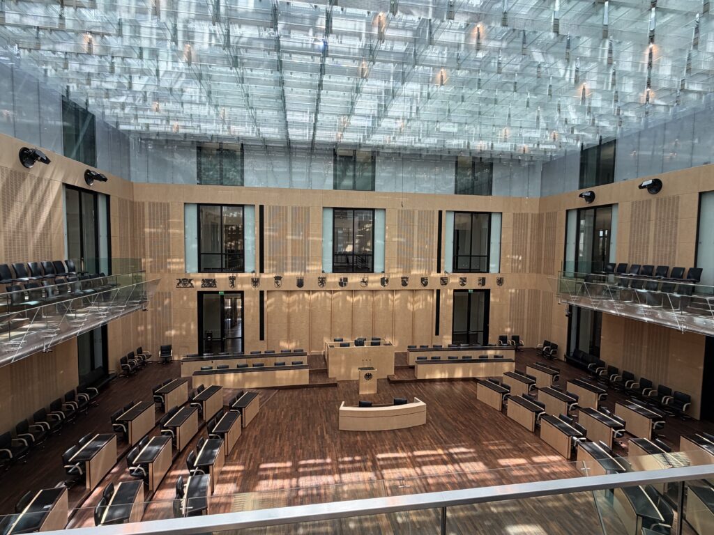 Leerer Plenarsaal mit Holzbestuhlung, mittigem Rednerpult, Zuschauerbalkonen und reflektierender Glasdecke.