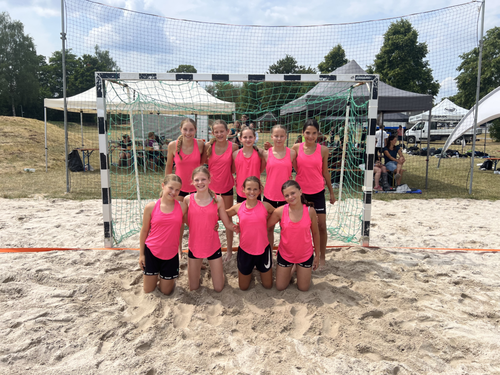 Mädchenhandballteam in leuchtend pinken Trikots, kniend und stehend vor einem Tor auf Sandplatz