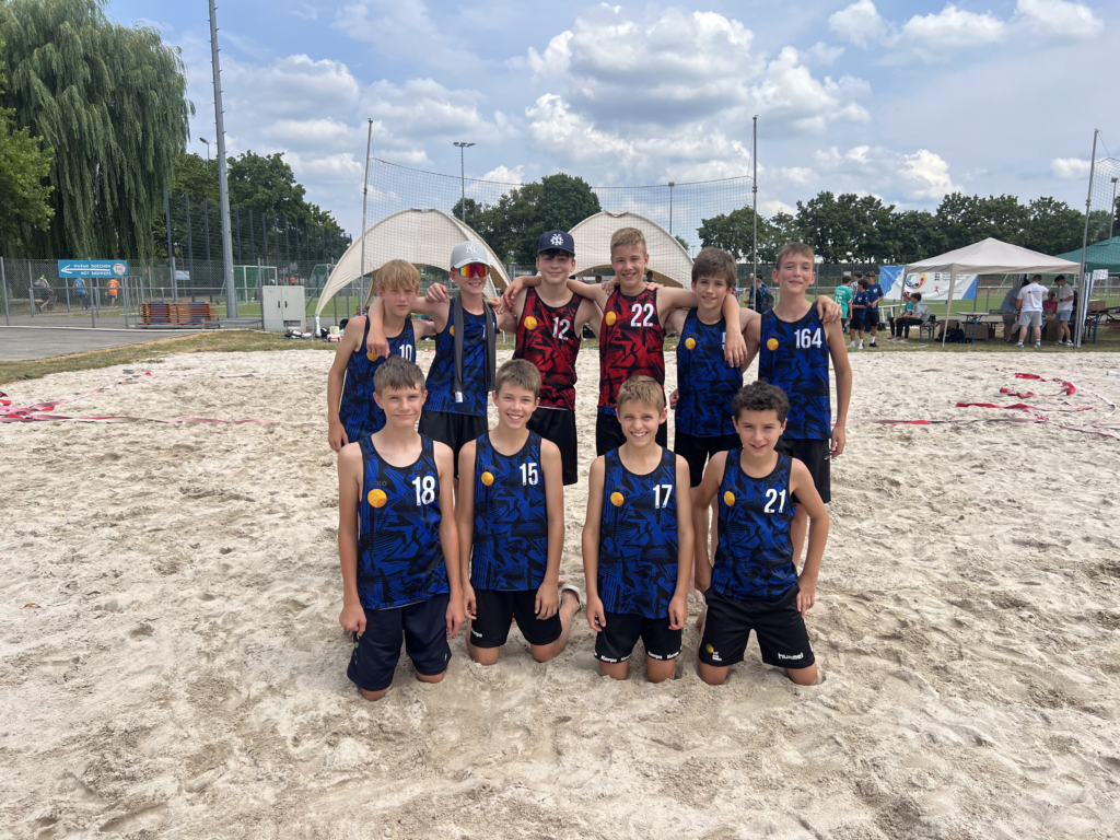 Jugendliches Beachvolleyball-Team posiert im Sand vor Netz und Turnierzelt, Jungen in Trikots lächelnd.