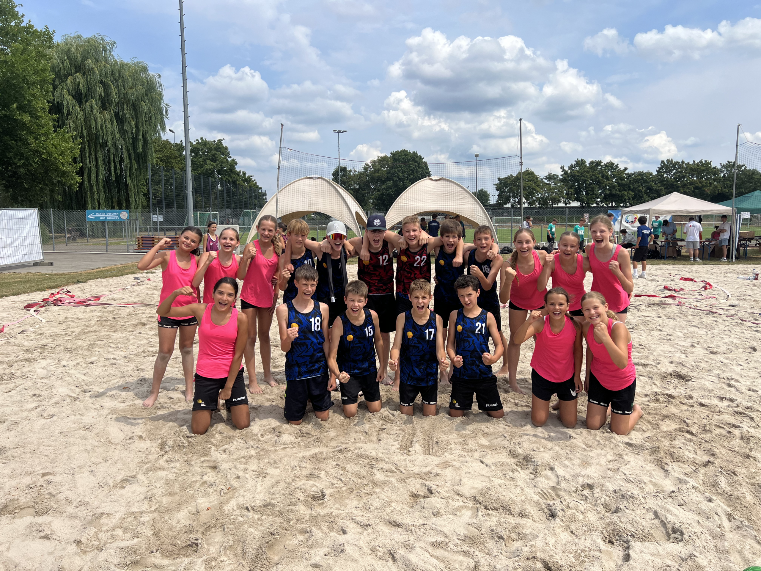 Kinder in pinken Trikots und blauen Jerseys jubeln zusammen auf einem Beachvolleyballfeld vor Zelten.