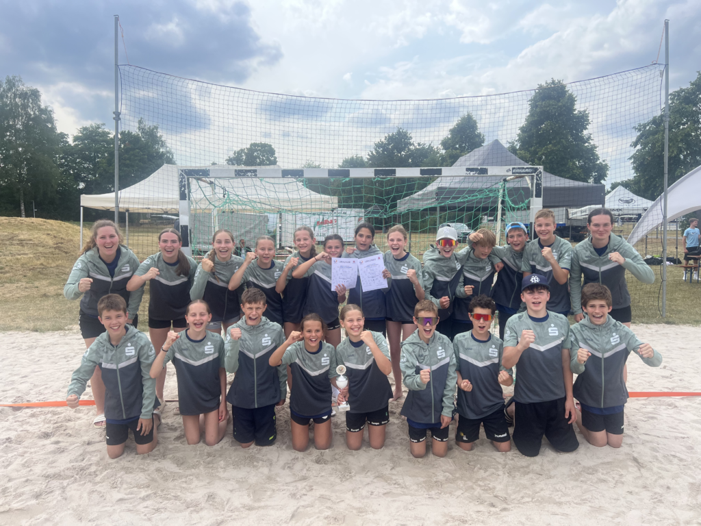 Junges Handballteam jubelt vor Tornetz am Sandplatz, einige halten Urkunden und einen Pokal.