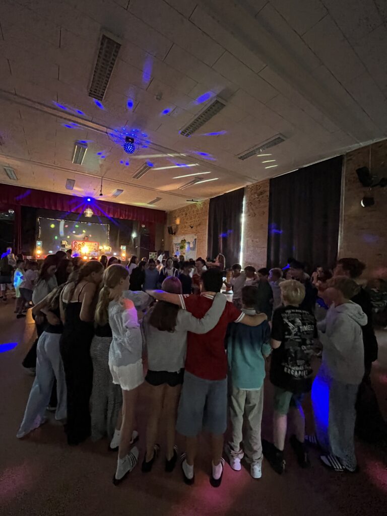 Jugendliche Arm in Arm auf Tanzfläche in dunkler Aula, bunte Diskolichter und Bühne im Hintergrund.