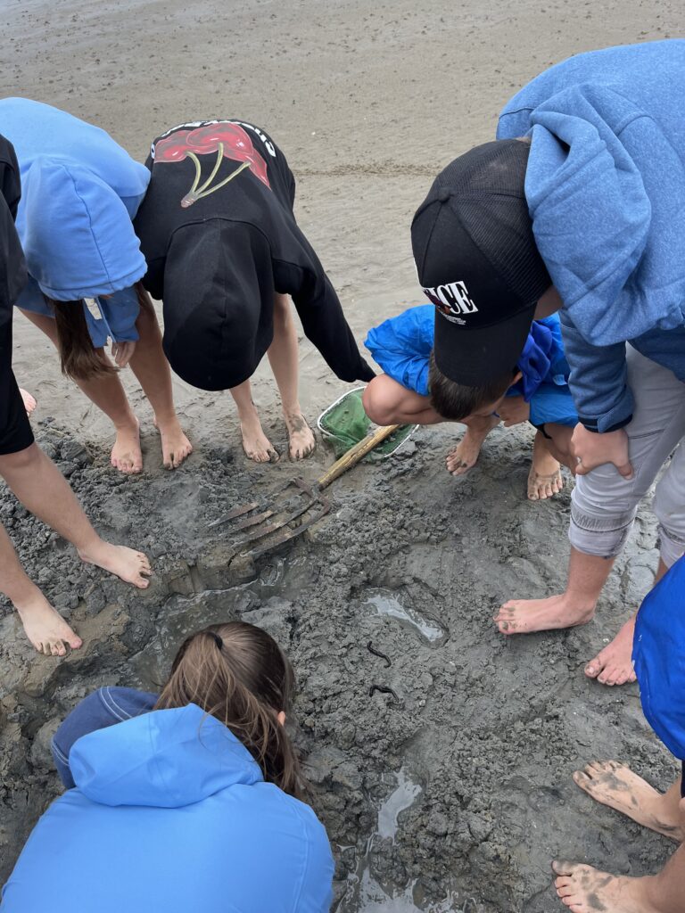 Mehrere barfüßige Kinder in Kapuzenpullis beugen sich über einen schlammigen Strand und beobachten kleine Würmer im Matsch.