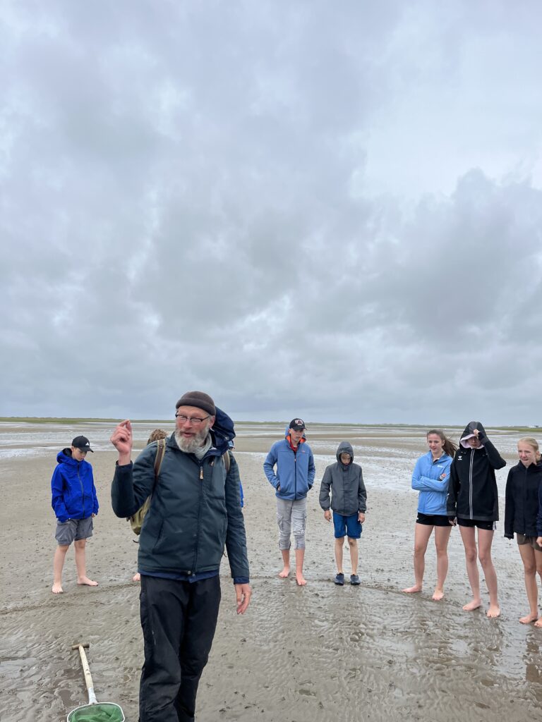 Mann mit Mütze erklärt einer Gruppe barfuß im Watt; grauer Himmel und Netz im Sand.