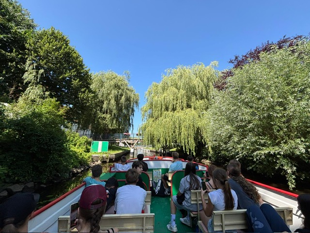 Passagiere auf Ausflugsboot fahren durch schmalen, baumgesäumten Kanal mit hängenden Weiden unter klarem blauem Himmel.