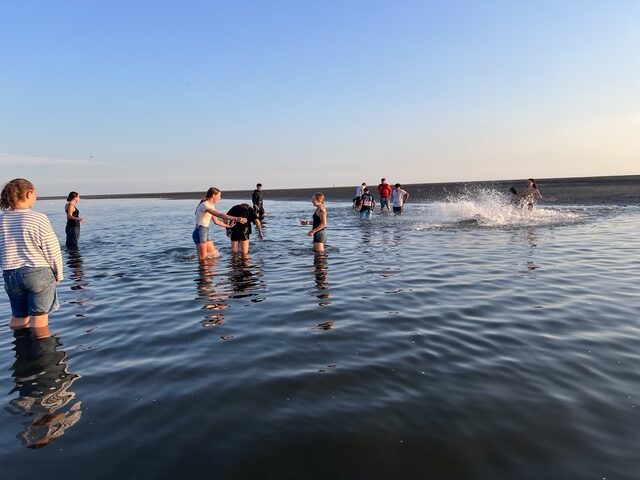 Gruppe von Menschen watet im flachen Meer bei Sonnenuntergang; einige spielen und spritzen Wasser.