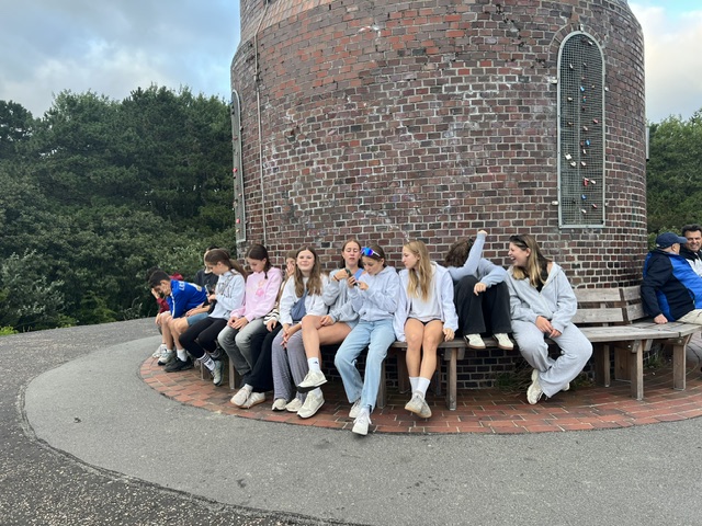 Mehrere Jugendliche sitzen dicht auf einer runden Bank um einen Backsteinturm, lässige Kleidung, Bäume im Hintergrund.