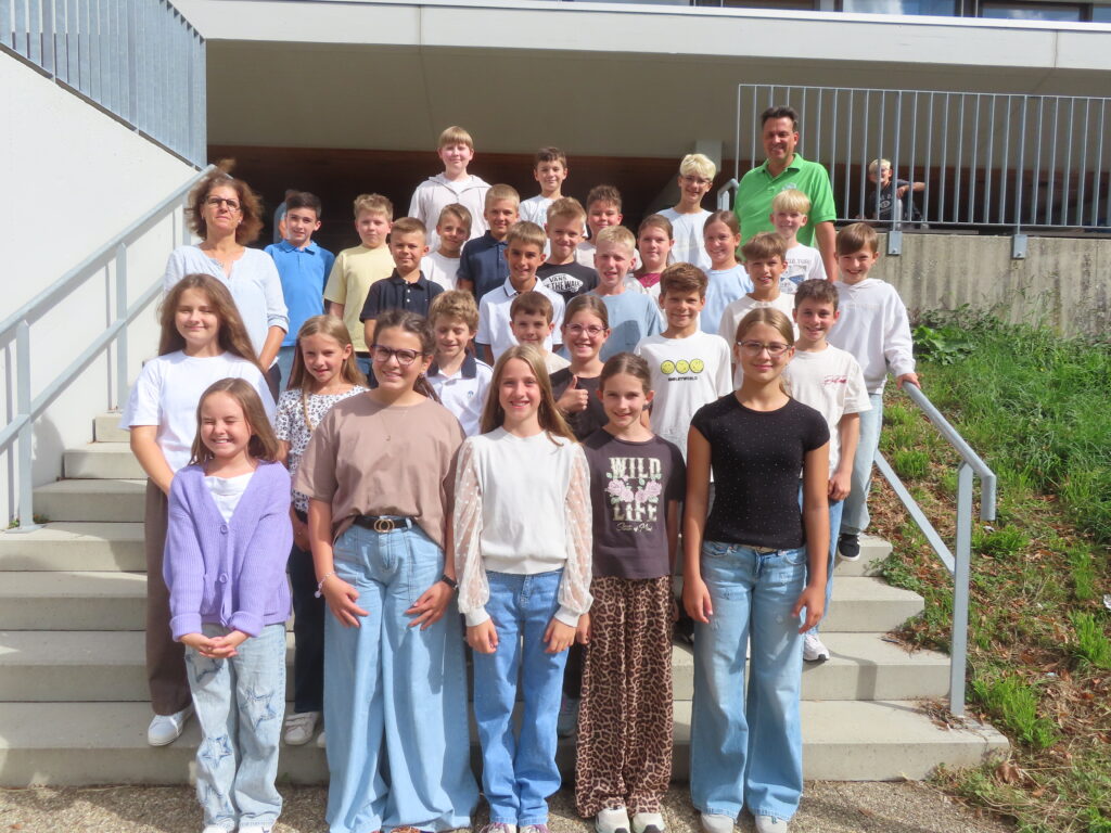 Schulklasse mit etwa 30 Kindern und zwei Lehrkräften auf Außentreppe, Gruppenfoto, freundlich lächelnd.