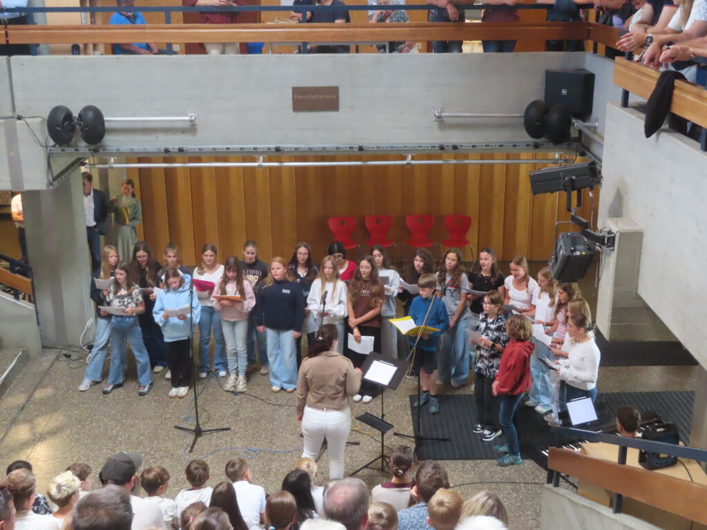 Schülerchor in Schul-Aula, Dirigentin leitet Jugendliche, Publikum sitzt auf Treppenstufen und Balkon.