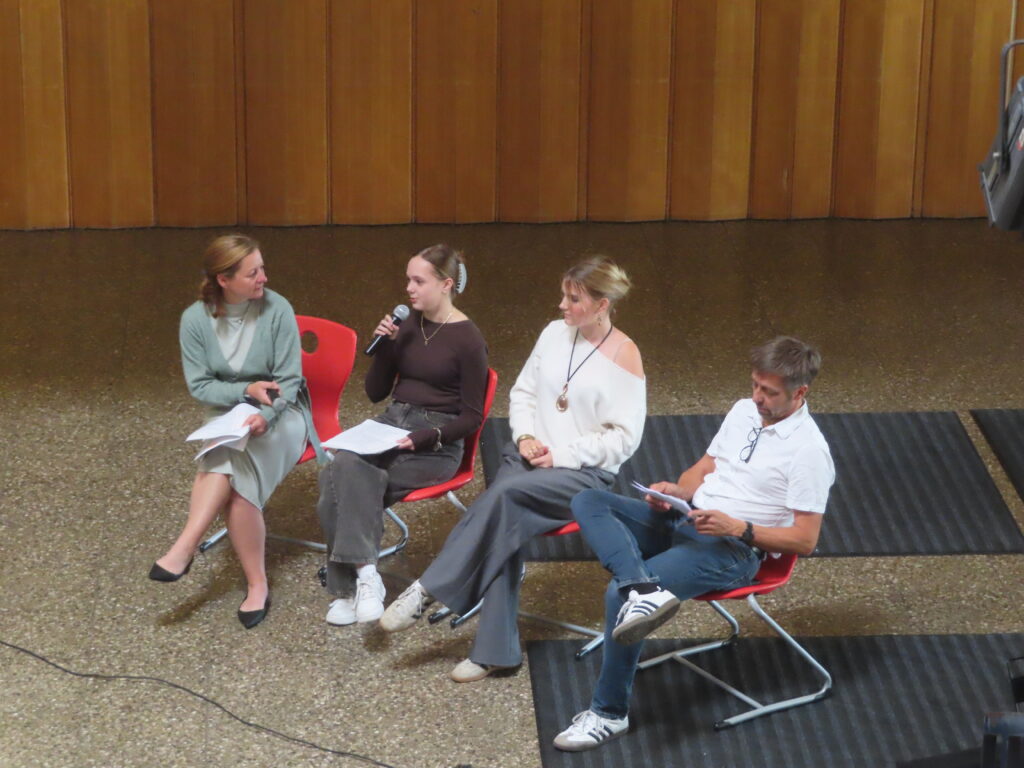 Vier Personen sitzen auf Stühlen bei einer Diskussion; junge Frau spricht ins Mikrofon, andere halten Unterlagen.