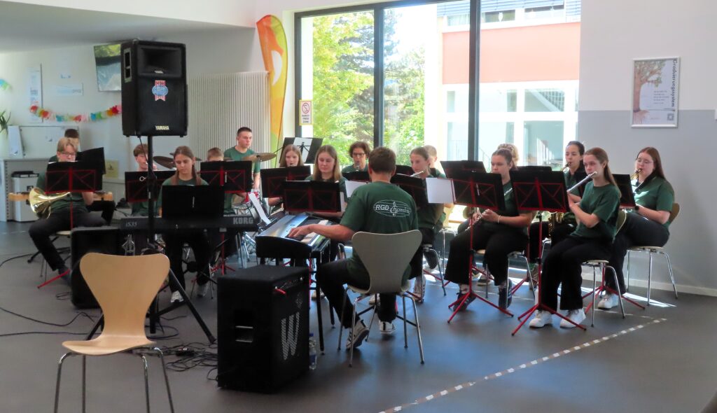 Jugendblasorchester in grünen T‑Shirts bei Probe in hellem Raum, mit Flöten, Saxophonen, Keyboard und Notenständern