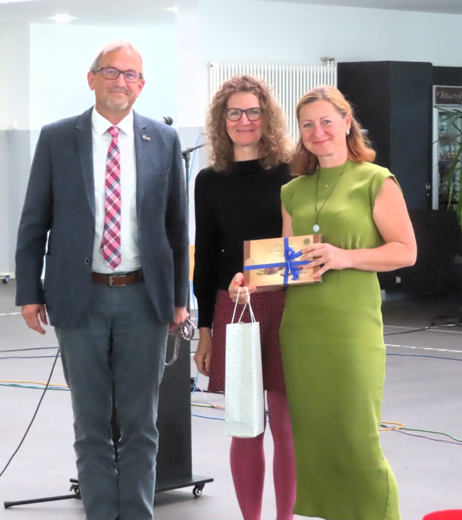 Drei lächelnde Erwachsene bei einer Ehrung; Frau in grün hält Geschenk mit blauem Band, neben ihr zwei Personen