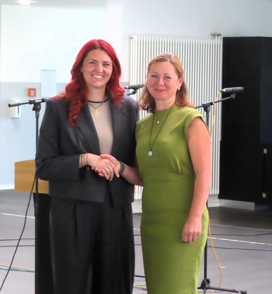 Zwei lächelnde Frauen geben sich die Hand; links rote Haare und dunkler Blazer, rechts grünes Kleid; Mikrofone im Hintergrund.