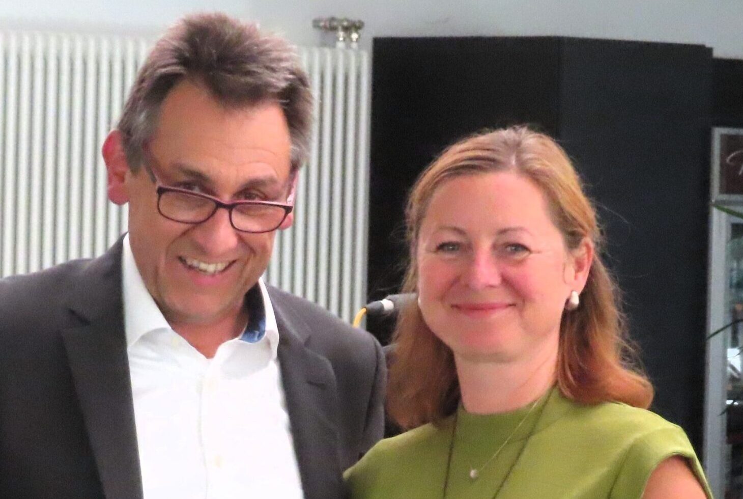 Zwei lächelnde Personen bei Veranstaltung, Mann mit Brille im Sakko hält gelbes Band, Frau in grünem Kleid
