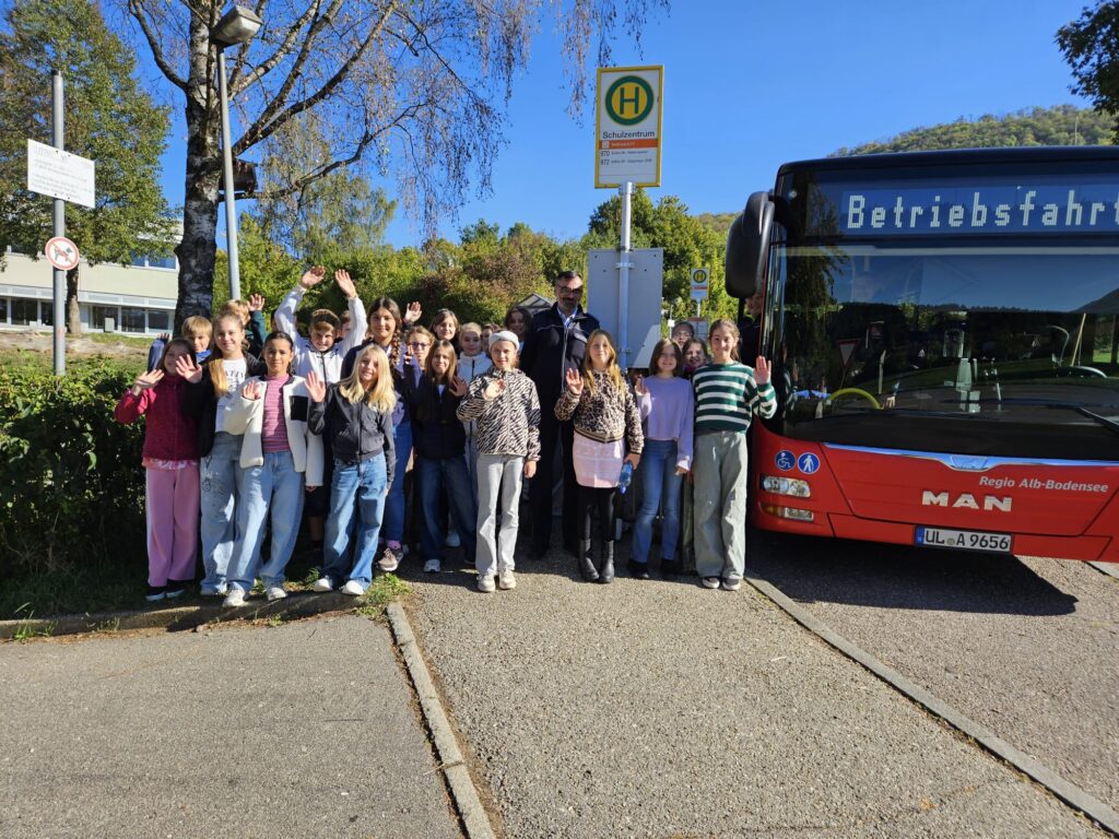Schulklasse mit Busfahrer winkt an Haltestelle vor rotem Bus, Anzeige Betriebsfahrt.