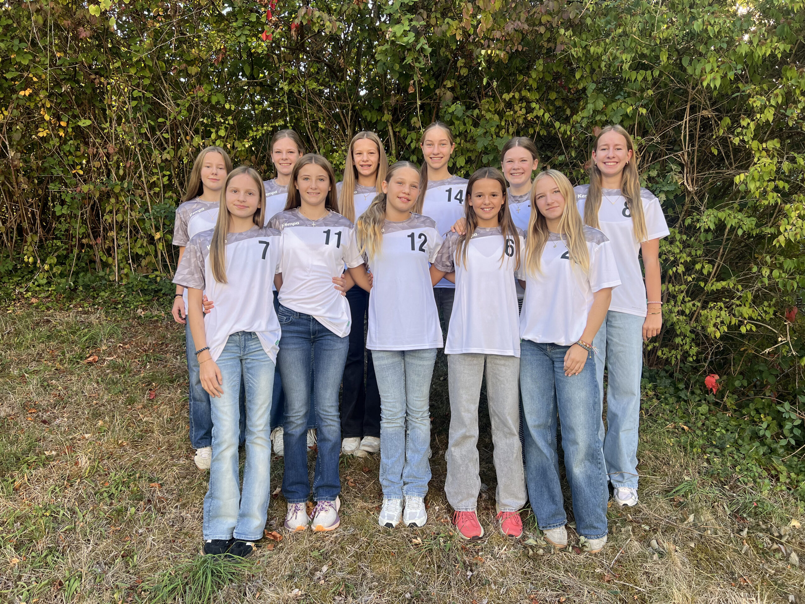 Elf lächelnde Mädchen in weißen Trikots mit Nummern stehen vor einer grünen Hecke, Teamfoto im Freien
