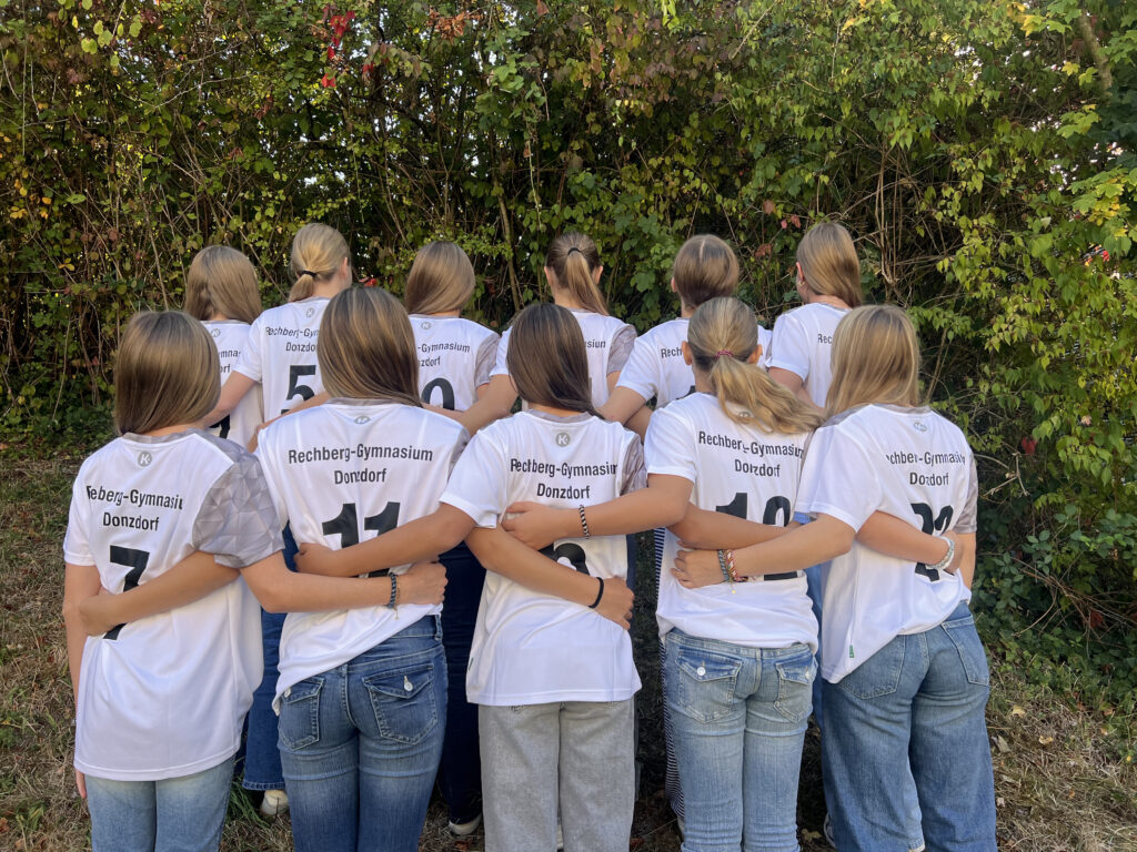 Gruppe Jugendlicher in weißen Trikots mit Rechberg-Gymnasium Donzdorf, Rücken zur Kamera, Arme umschlungen vor Gebüsch.