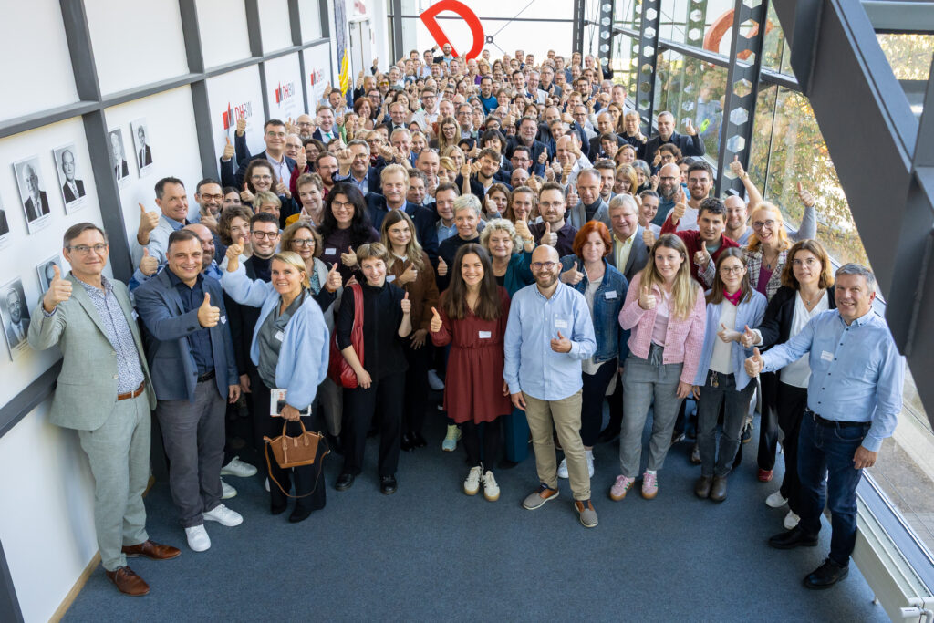 Große Gruppe lächelnder Menschen in hellem Glasgang, Gruppenfoto bei Veranstaltung; alle zeigen Daumen hoch