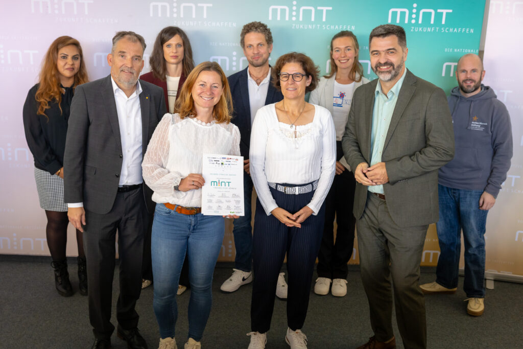 Neun Personen vor MINT‑Banner, zwei Frauen vorn halten eine Auszeichnung MINT‑freundliche Schule und lächeln