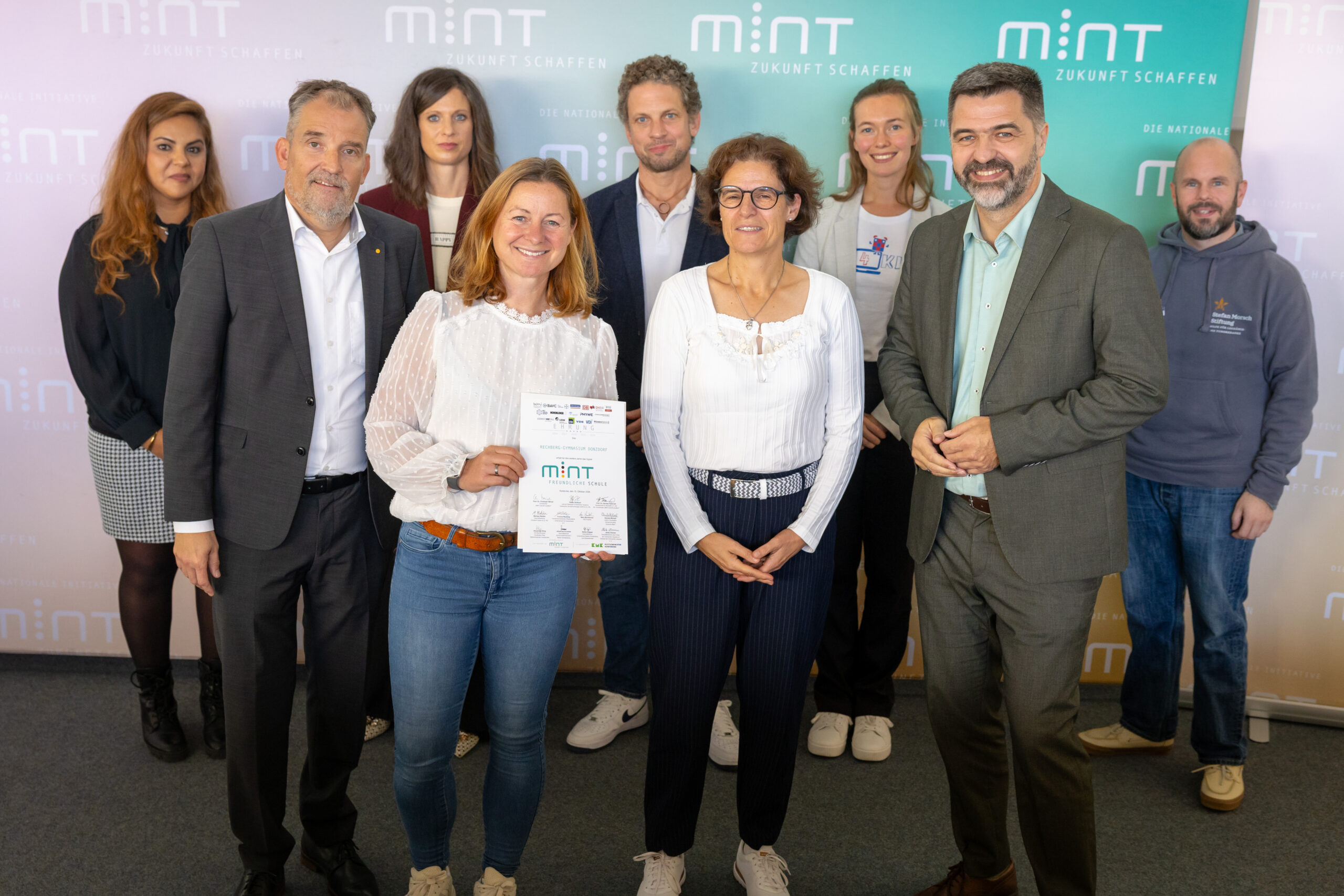 Neun Personen vor MINT‑Banner, zwei Frauen vorn halten eine Auszeichnung MINT‑freundliche Schule und lächeln